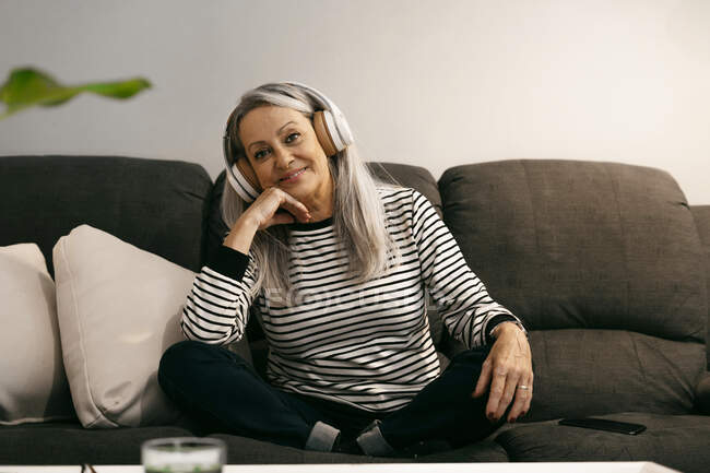 Seniorin mit Kopfhörer lächelt, während sie zu Hause mit der Hand am Kinn sitzt — Stockfoto