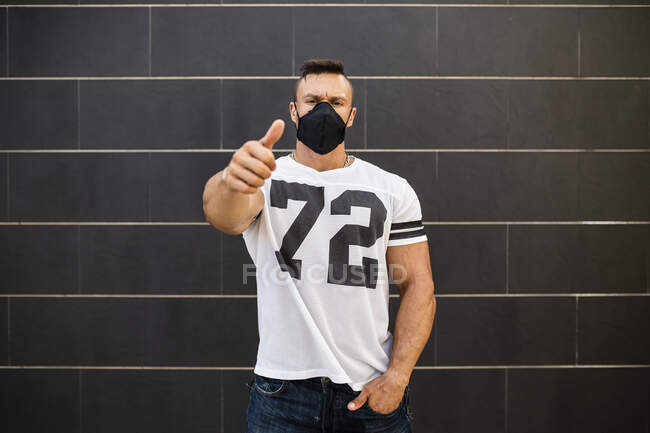Macho homme portant un masque facial montrant pouces levés tout en se tenant contre le mur — Photo de stock