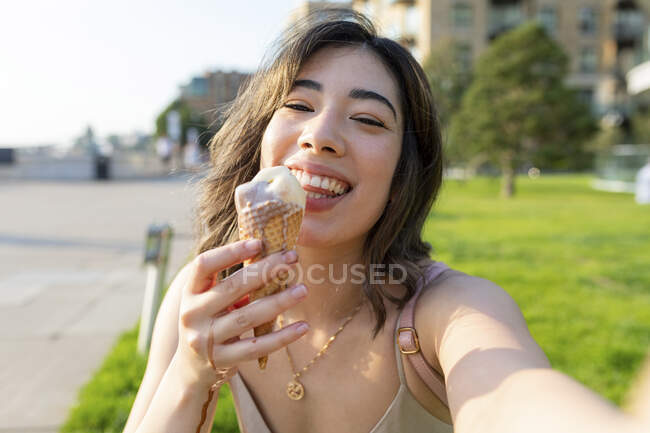 Smiling woman eating ice cream while taking selfie in public park on sunny day — Photo de stock