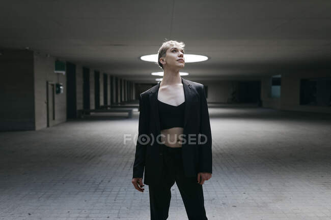 Thoughtful trans young man wearing black suit standing on floor in basement — Stock Photo