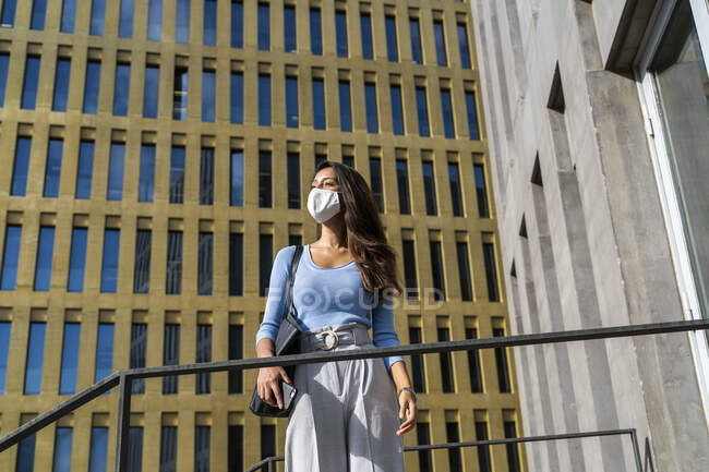Joven mujer de negocios con máscara facial mirando hacia otro lado mientras está de pie contra el edificio de oficinas durante el brote de coronavirus - foto de stock