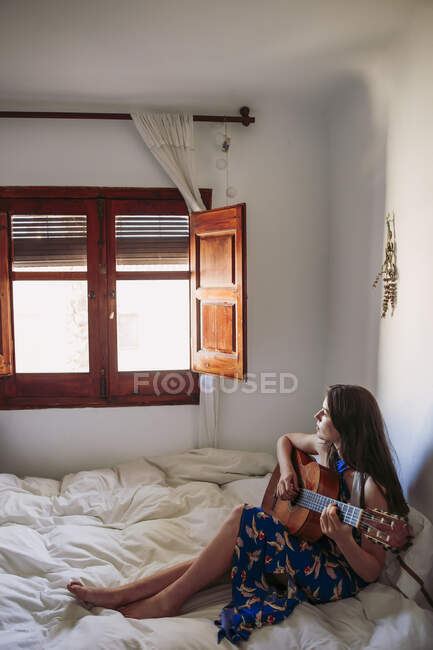 Жінка дивиться через вікно, практикуючи гітару в спальні вдома — стокове фото