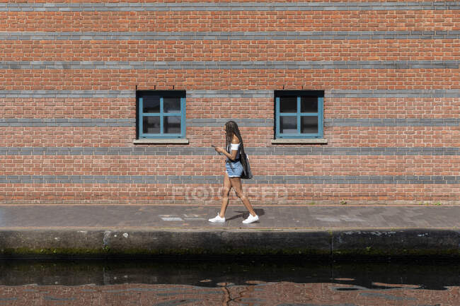 Жінка використовує телефон під час ходьби на тротуарі навпроти цегляної стіни — стокове фото