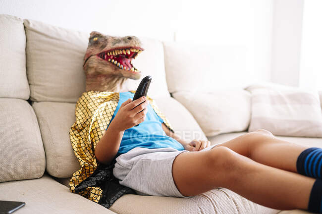 Хлопчик у масці динозавра дивиться телевізор, відпочиваючи на дивані вдома. — стокове фото