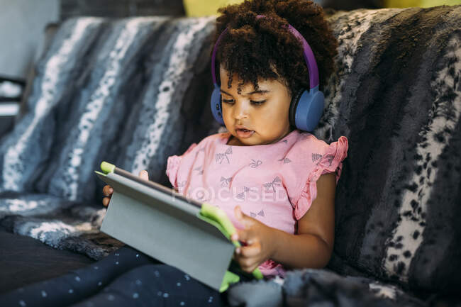 Baby Mädchen mit lockigem Haar mit digitalem Tablet zu Hause auf dem Sofa sitzend — Stockfoto