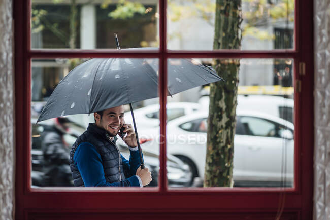Hombre feliz con paraguas hablando en el teléfono inteligente mientras mira a la ventana durante la temporada de lluvias - foto de stock