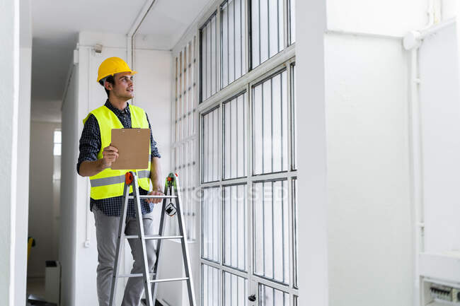 Homme portant casque de travail et veste debout sur l'échelle par fenêtre sur le chantier de construction — Photo de stock