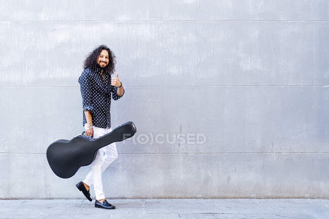 Smiling man with guitar showing thumps up while walking against gray wall — Stock Photo