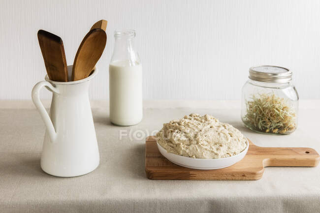 Jarra con cuchara y espátula de madera, botella de leche, frasco de brotes y masa en tabla de cortar - foto de stock