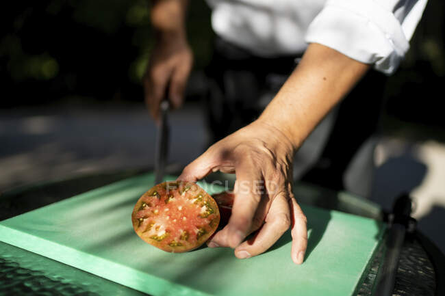 Gros plan d'un chef masculin tenant une tranche de tomate sur une planche à découper dans un verger — Photo de stock