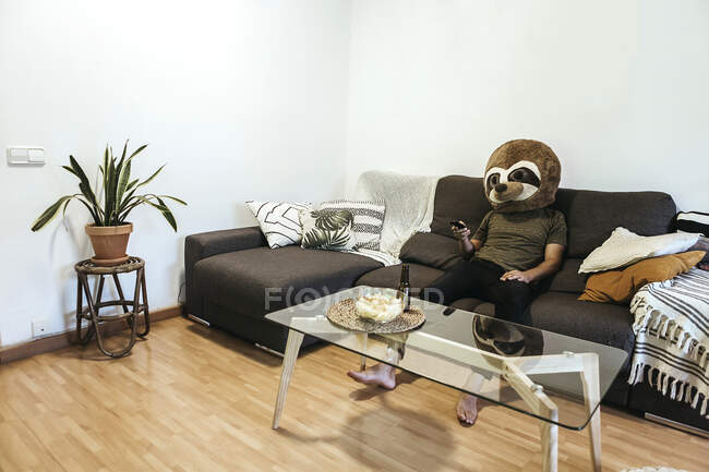Hombre con máscara de oso de peluche viendo la televisión mientras se relaja en el sofá en la sala de estar - foto de stock