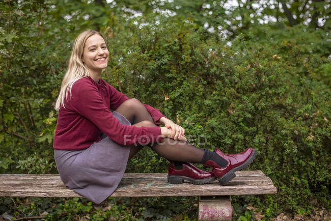 Mujer rubia sonriente sentada en el banco por las plantas en el parque durante el otoño - foto de stock