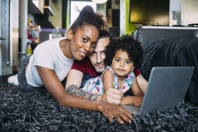 Los padres con hija usando el ordenador portátil mientras están acostados en la alfombra en casa - foto de stock