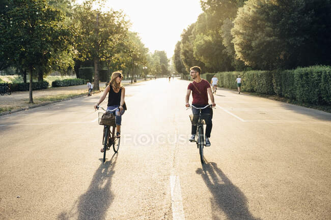 Щаслива пара катається на велосипеді по дорозі серед дерев у парку під час заходу сонця — стокове фото