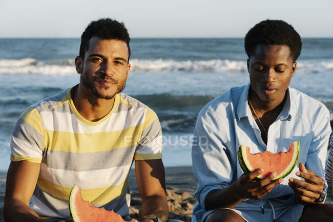 Jóvenes amigos comiendo sandía sentados en la playa - foto de stock