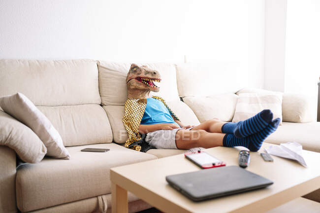 Ragazzo che indossa maschera di dinosauro rilassante sul divano di casa — Foto stock