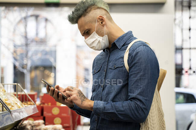 Stylish man wearing face mask using smart phone in supermarket — Stock Photo