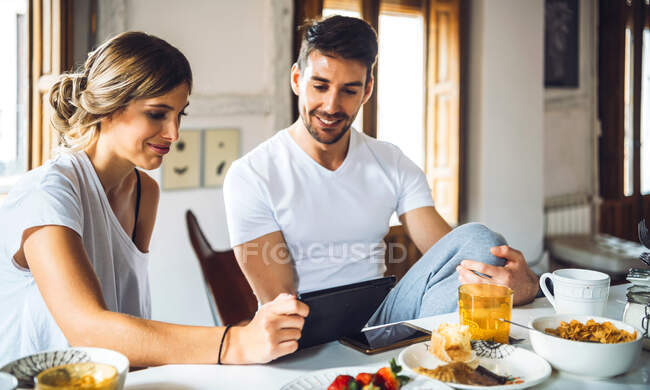 Jovem casal assistindo tablet enquanto toma café da manhã em casa — Fotografia de Stock