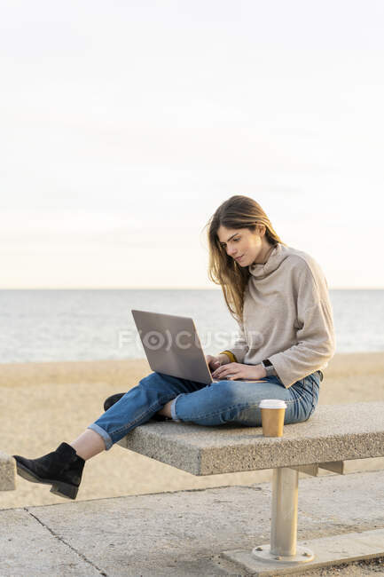 Belle femme utilisant un ordinateur portable tout en étant assis avec une tasse jetable sur le banc à la promenade par la mer contre le ciel — Photo de stock