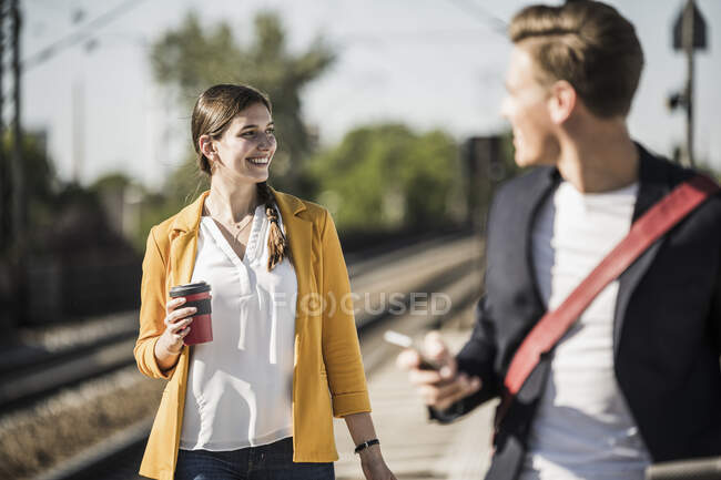 Donna sorridente con tazza di caffè riutilizzabile guardando lontano mentre camminava sulla piattaforma della stazione ferroviaria — Foto stock
