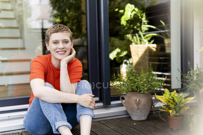 Улыбчивая красивая женщина с рукой на подбородке, сидящая напротив дома во дворе — стоковое фото