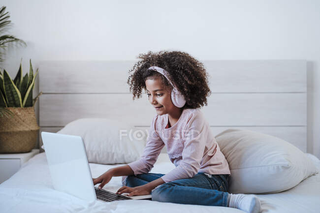 Мила дівчина користується ноутбуком і носить навушники, сидячи вдома у спальні. — стокове фото