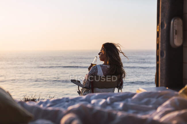Young woman drinking white wine while sitting on chair by camper van at beach during sunset — Stock Photo