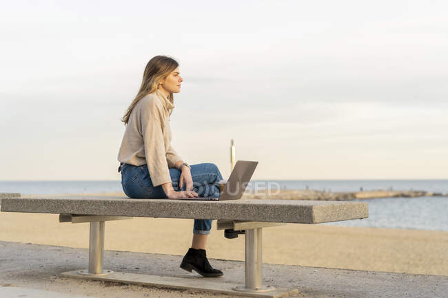 Nachdenkliche Frau schaut weg, während sie bei Sonnenuntergang mit Laptop auf Bank an der Strandpromenade gegen den Himmel sitzt — Stockfoto