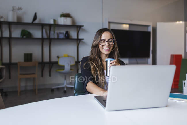 Femme d'affaires souriante buvant du café tout en utilisant un ordinateur portable au bureau — Photo de stock