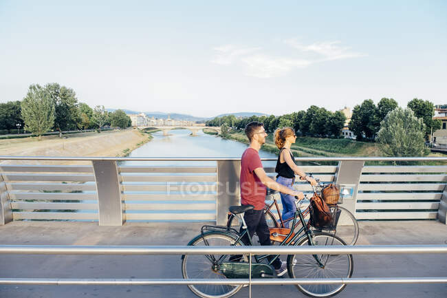 Casal com bicicletas andando na ponte sobre o rio contra o céu — Fotografia de Stock