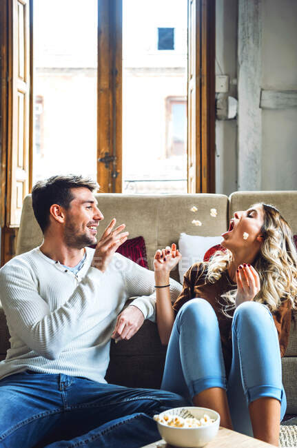 Jovem casal alegre sentado em casa e brincando com pipocas — Fotografia de Stock