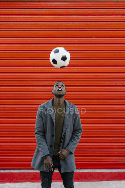 Jeune homme jouant avec le football tout en se tenant contre le mur rouge — Photo de stock