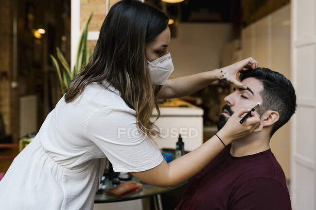 Young female stylist applying make-up to groom in salon during coronavirus — Stock Photo