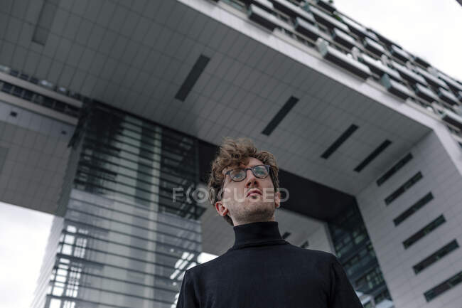 Schwerwiegender junger Mann schaut weg, während er gegen modernes Gebäude in der Stadt steht — Stockfoto
