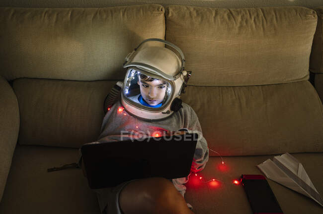 Boy wearing space helmet using laptop while sitting with illuminated lighting equipment on sofa at home — Stock Photo