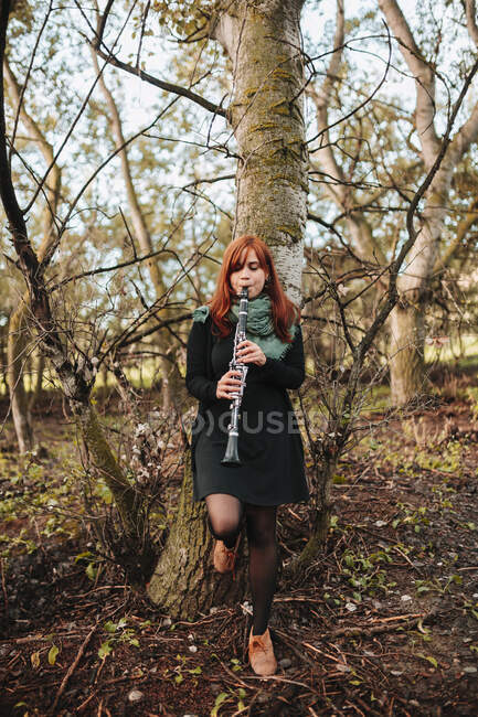 Bella giovane donna che pratica il clarinetto mentre si appoggia sul tronco nudo dell'albero nella foresta — Foto stock