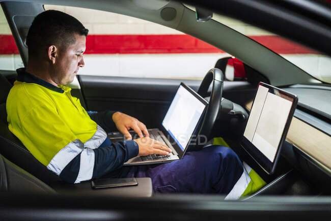 Male technician programming on laptop while sitting in electric car — Stock Photo