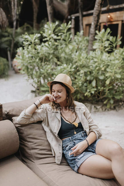 Усміхнена жінка з капелюхом сидить на дивані на відкритому повітрі — стокове фото