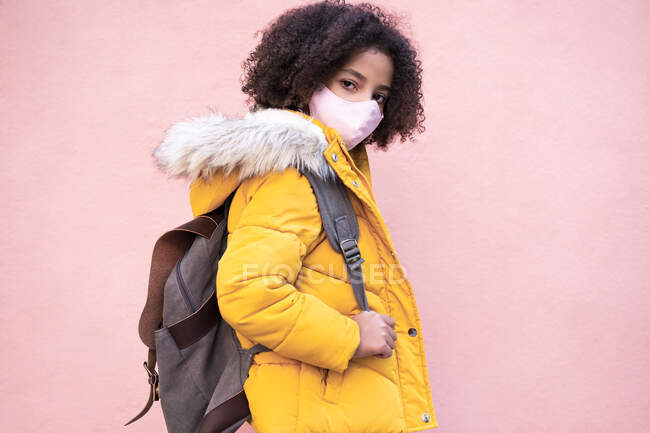 Chica con mascarilla que lleva la mochila mientras está de pie contra la pared rosa — Stock Photo