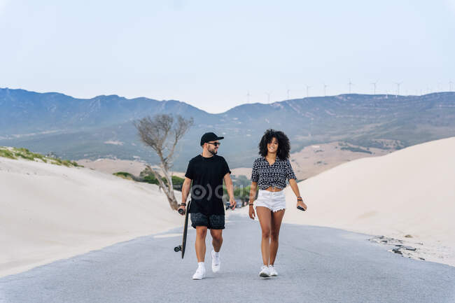 Smiling multi-ethnic couple walking on road amidst sand dunes against clear sky — Stock Photo