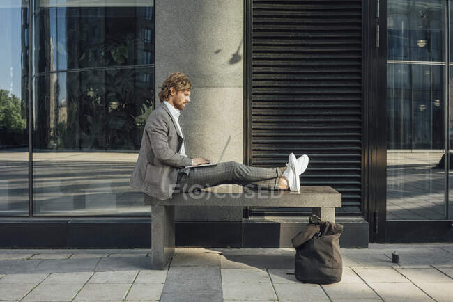 Мужчина-профессионал с помощью ноутбука, сидя на скамейке со скрещенными ногами в центре города — стоковое фото