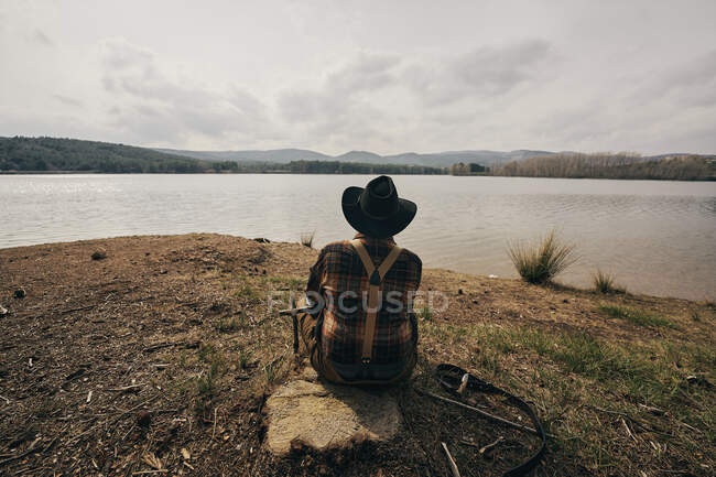 Bushcrafter sentado en el bosque mientras mira el lago durante el atardecer - foto de stock