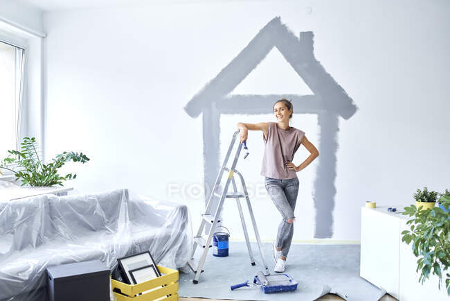 Femme souriante avec la main sur la hanche debout contre la maison peinte sur le mur à la maison — Photo de stock