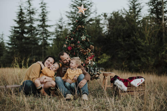 Família sorridente sentada à beira da árvore de Natal em terras gramadas no campo durante o pôr do sol — Fotografia de Stock