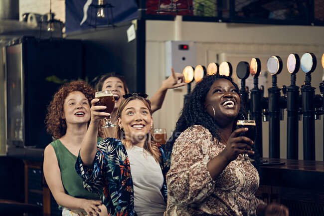 Emocionadas amigas sentadas en el mostrador en un pub viendo Tv - foto de stock