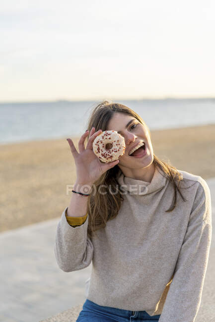 Веселая молодая женщина держит свежий пончик перед лицом, сидя на пляже во время заката — стоковое фото