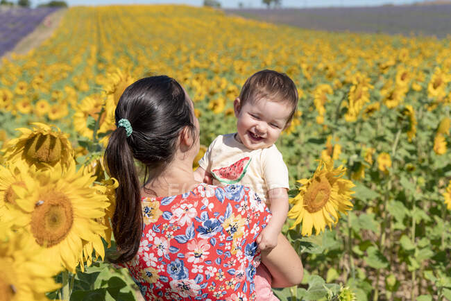 Madre llevando alegre hija en el campo de girasol durante el verano - foto de stock