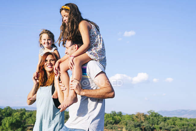 Padres felices llevando hijas en hombros contra el cielo durante el día soleado - foto de stock