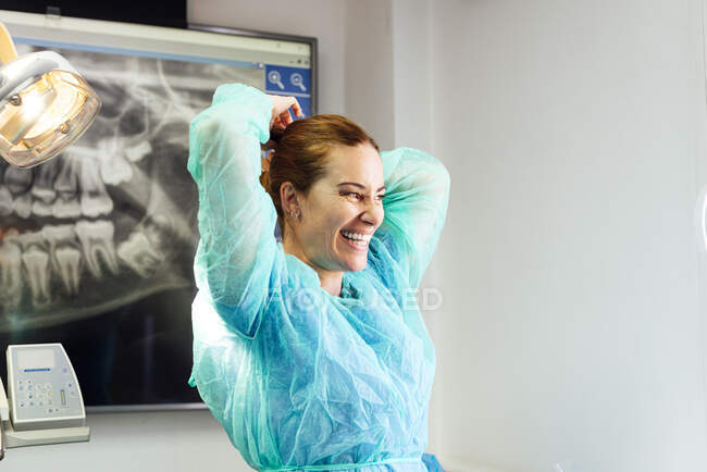 Lächelnder Zahnarzt im Schutzanzug, der im Stehen in der Klinik Haare bindet — Stockfoto