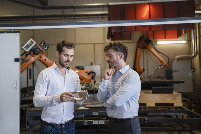Empresario con objeto de metal discutiendo con su colega mientras está de pie contra la máquina robótica en la fábrica - foto de stock